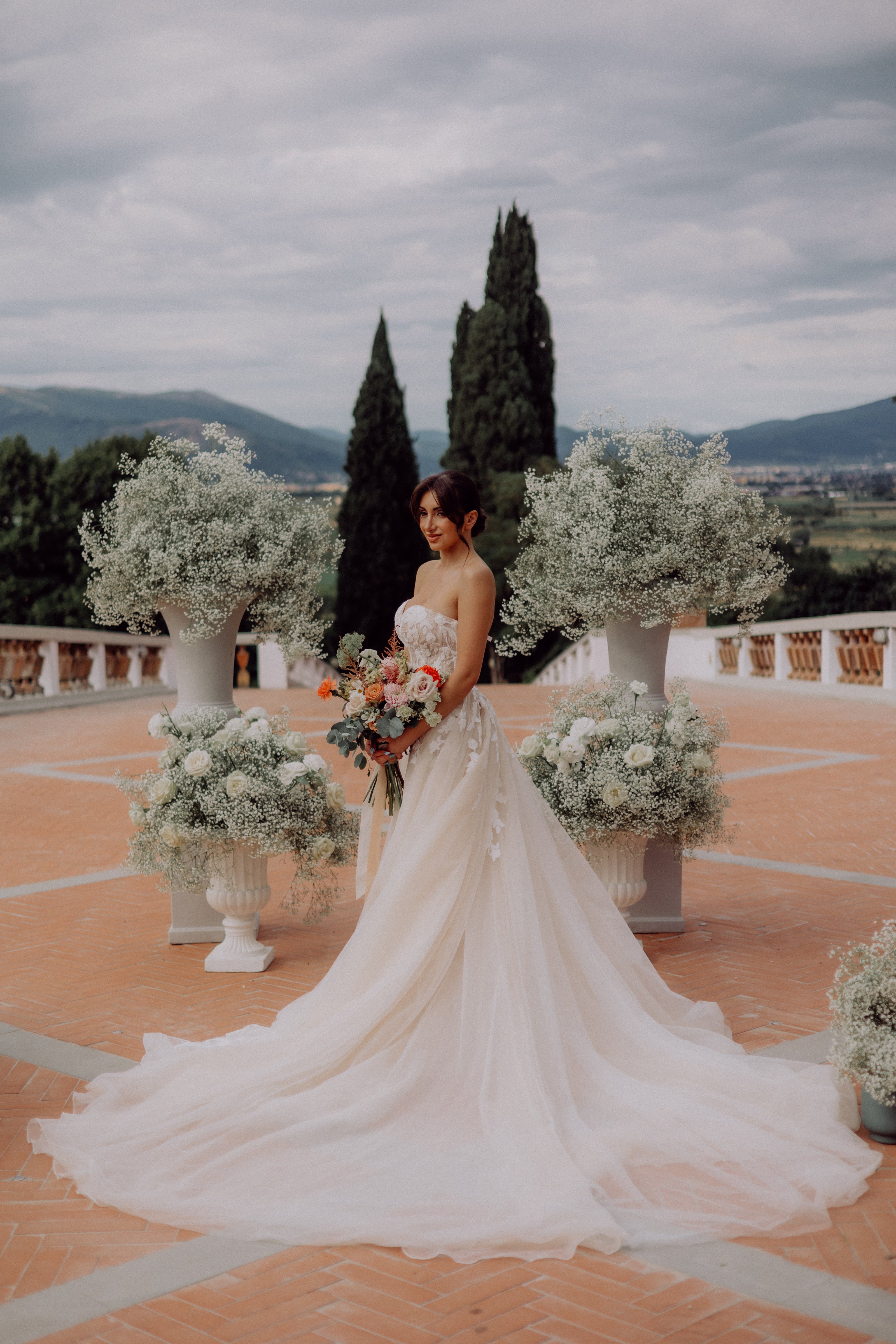 Bride standing at the aisle with over the top babies breath in vases and the Tuscany hills behind her in a gorgeous white dress that drapes over the floor
