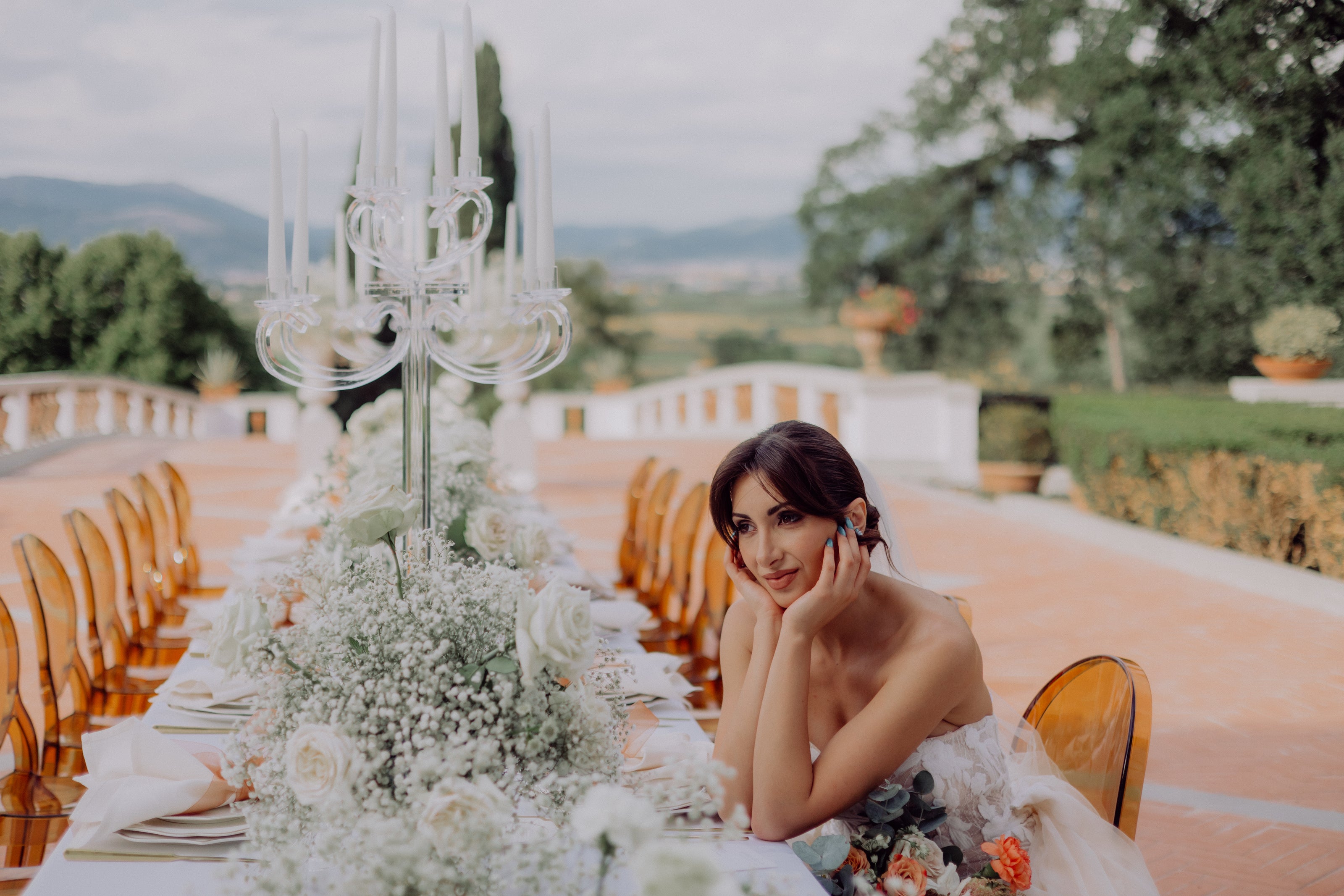 A Bride sitting at her wedding table looking happy and content roaming hills of Tuscany in Italy and table decorated in babies breath flowers and clear candelabras