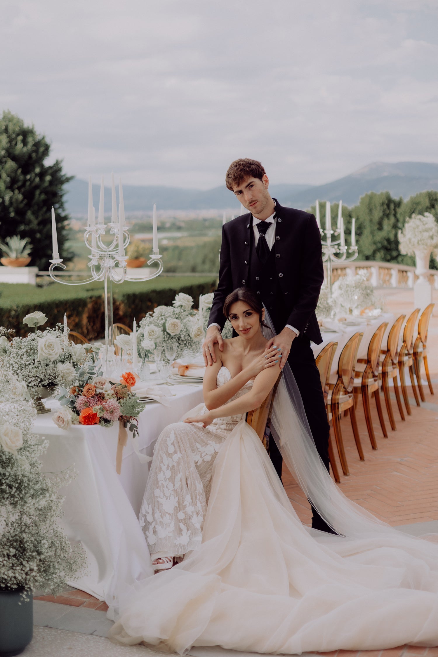 Bride and Groom at their long table wedding, portrait style showcasing the roaming plains of Tuscany Italy and ghost chairs, flowers and clear candelabras